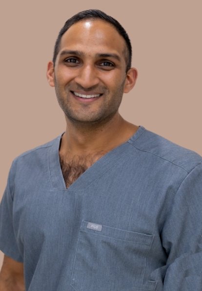 Ageless Smiles Dental Centre Smiling man in grey medical scrubs posing against a plain beige background.