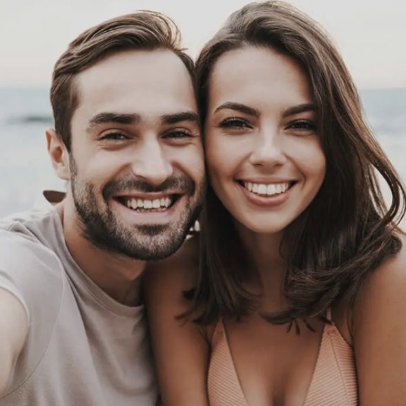 Ageless Smiles Dental Centre Smiling couple taking a close-up selfie at the beach, with the ocean visible in the blurred background.